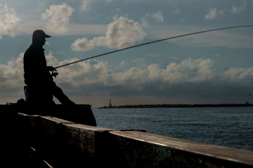 A man fishing off a sea wall, silhouetted against a cloudy sky